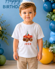 A young boy wearing a white t-shirt with a red truck graphic and the name " Josh" printed on it, standing in front of a blue birthday backdrop with balloons.