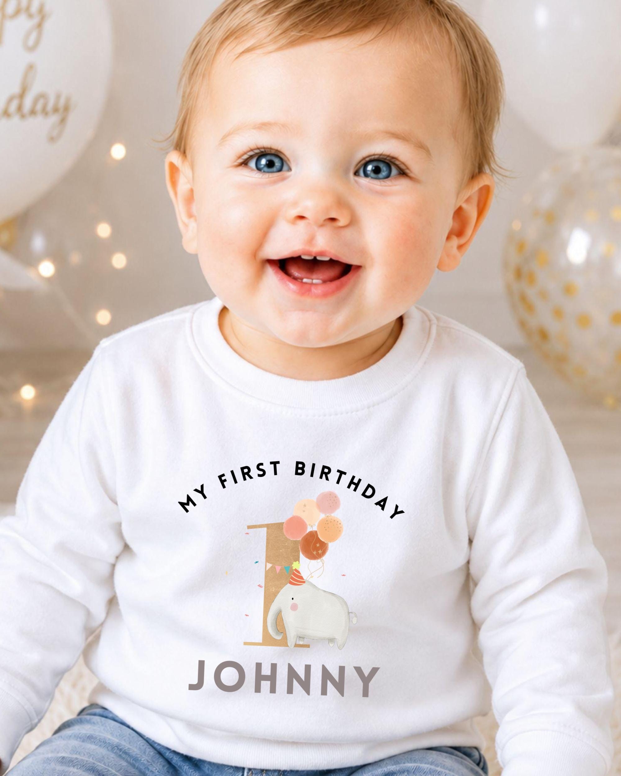 A smiling baby boy wearing a white shirt with a birthday message and an elephant graphic.