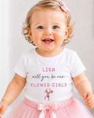 A smiling baby girl wearing a white t-shirt with text and a pink bow, standing in front of a white background.
