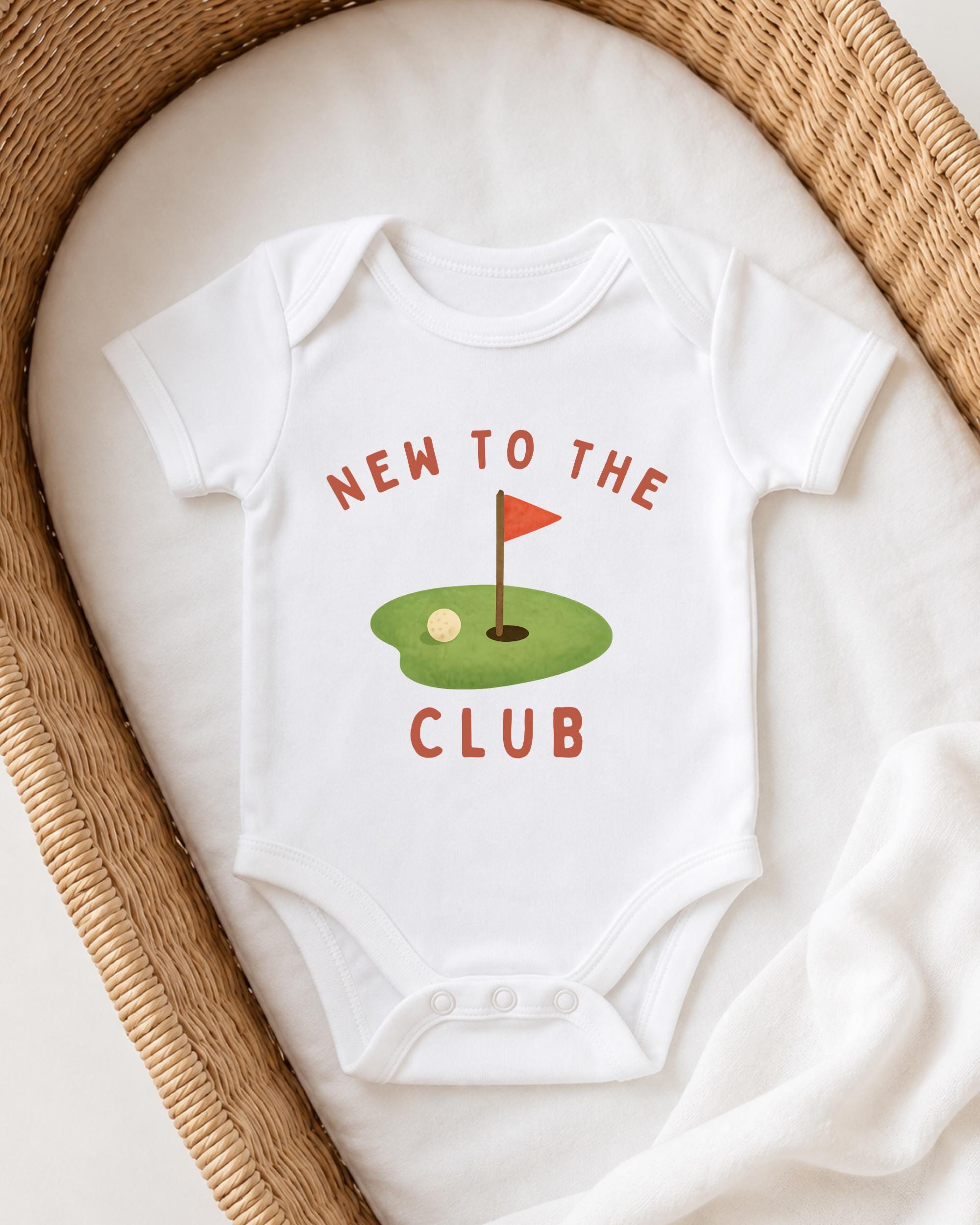 A white onesie with the text "NEW TO THE CLUB" printed on it, along with a golf hole graphic, is displayed on a white blanket.