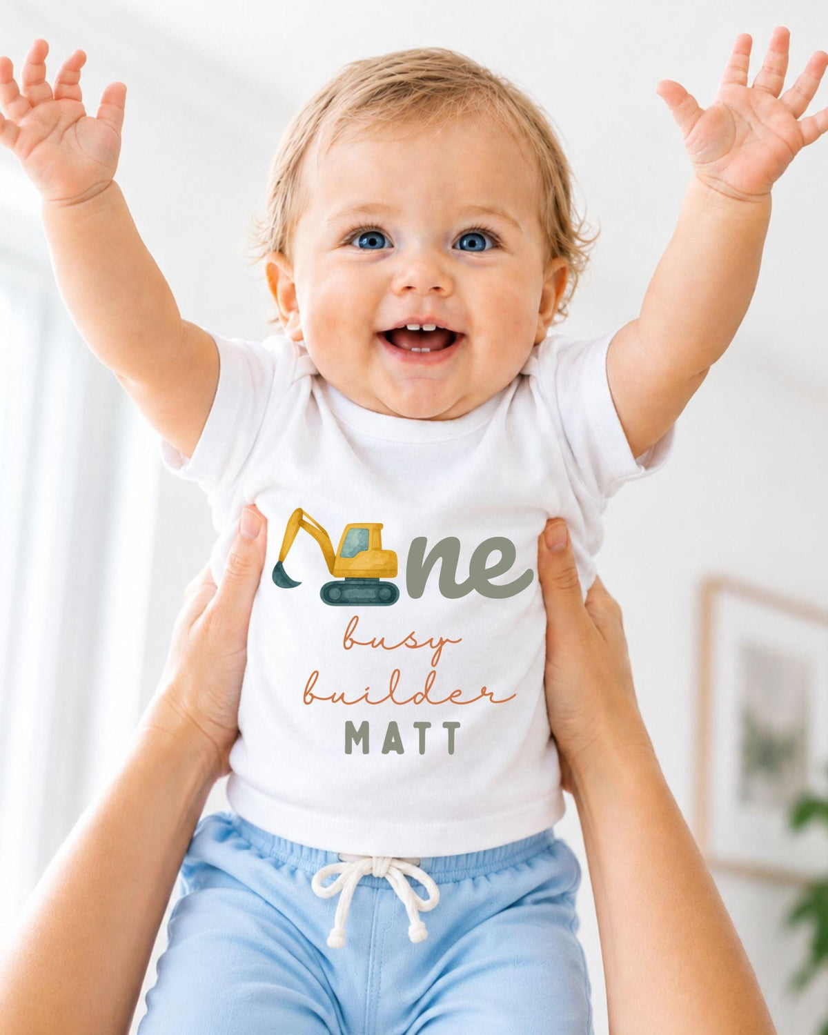A smiling baby wearing a white shirt with a construction vehicle graphic and the text "One busy builder Matt" is being held up by someone's arms.
