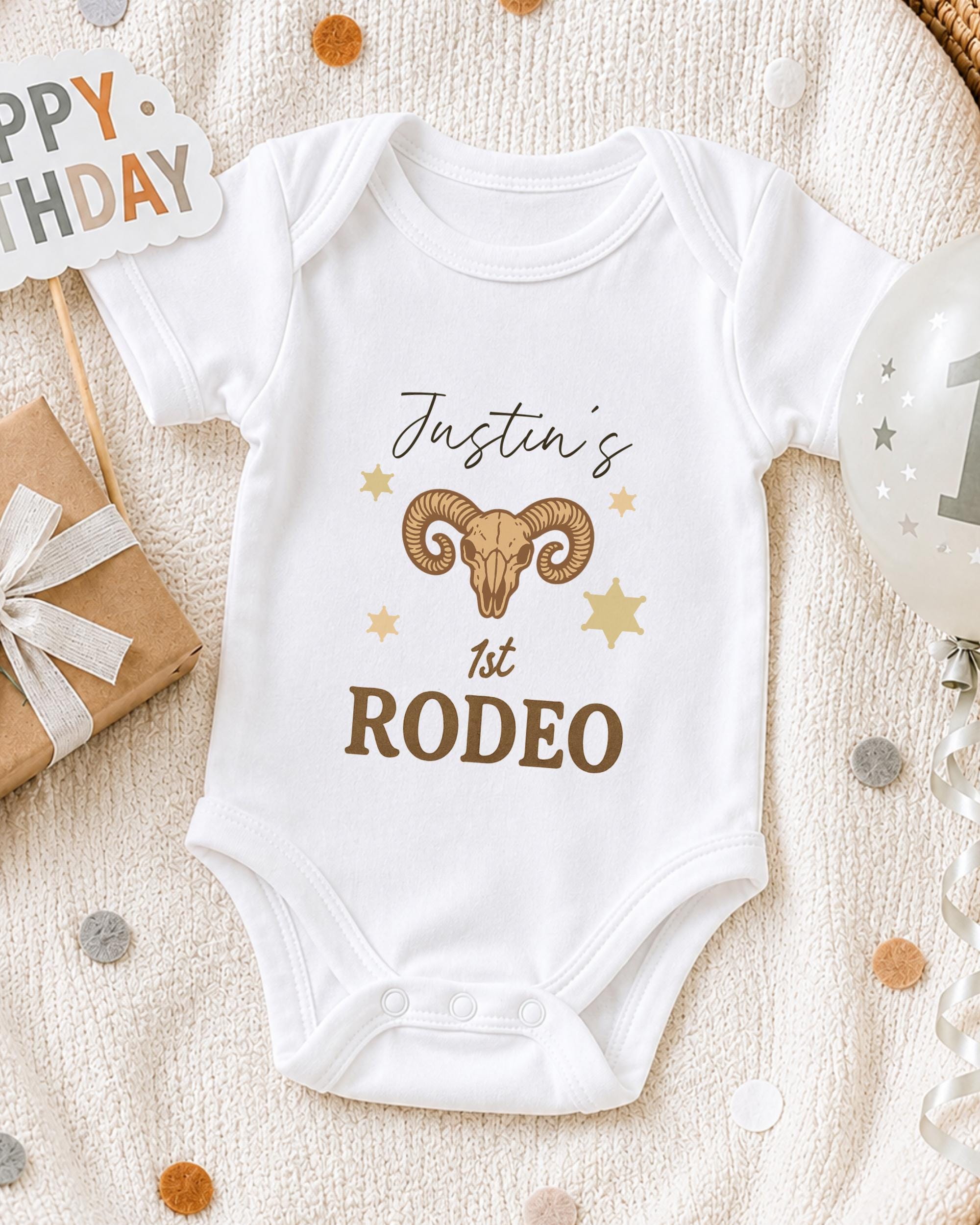 A white onesie with a graphic of a ram and the text "Justin's 1st Rodeo" printed on it, along with a "Happy Birthday" sign and a birthday balloon.