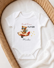 A white onesie with a cartoon image of a bear wearing a pilot's hat and flying an airplane, along with the text "First Time Flyer" and "Miki".