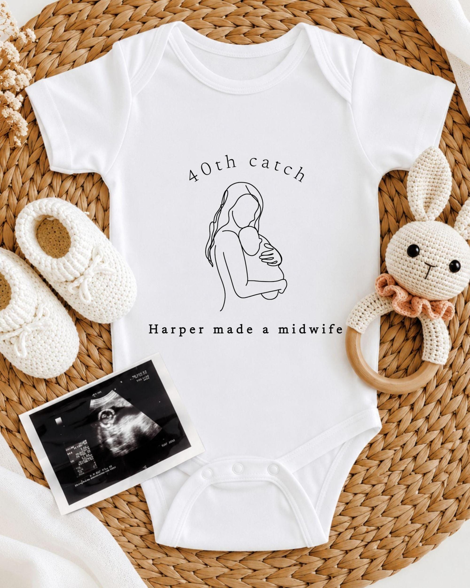 A white onesie with a graphic of a woman holding a baby, along with a pair of crocheted baby booties and a baby rattle, all arranged on a woven placemat.