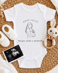 A white onesie with a graphic of a woman holding a baby, along with a pair of crocheted baby booties and a baby rattle, all arranged on a woven placemat.