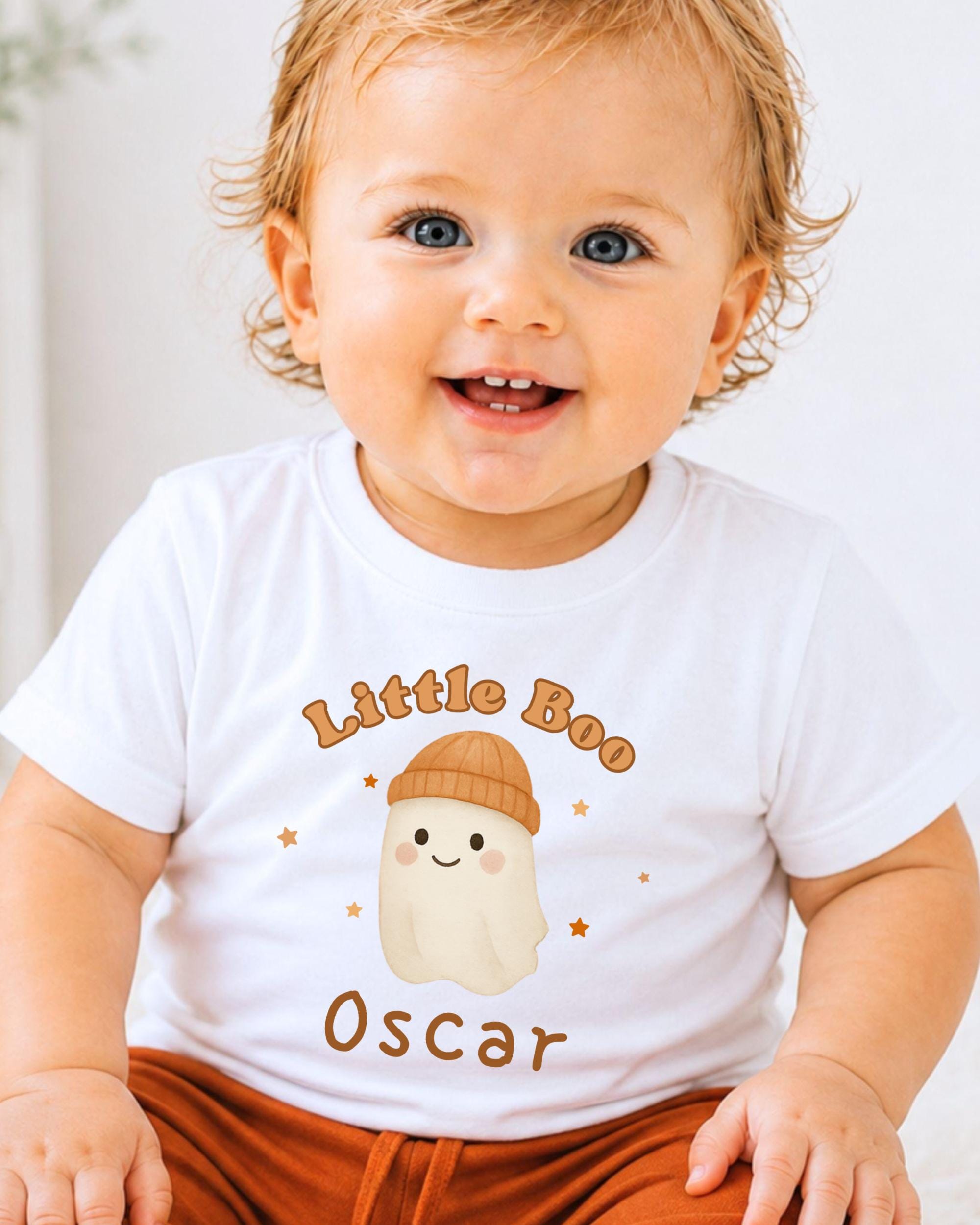 A smiling baby boy wearing a white t-shirt with a cartoon ghost and the text "Little Boo Oscar" printed on it.