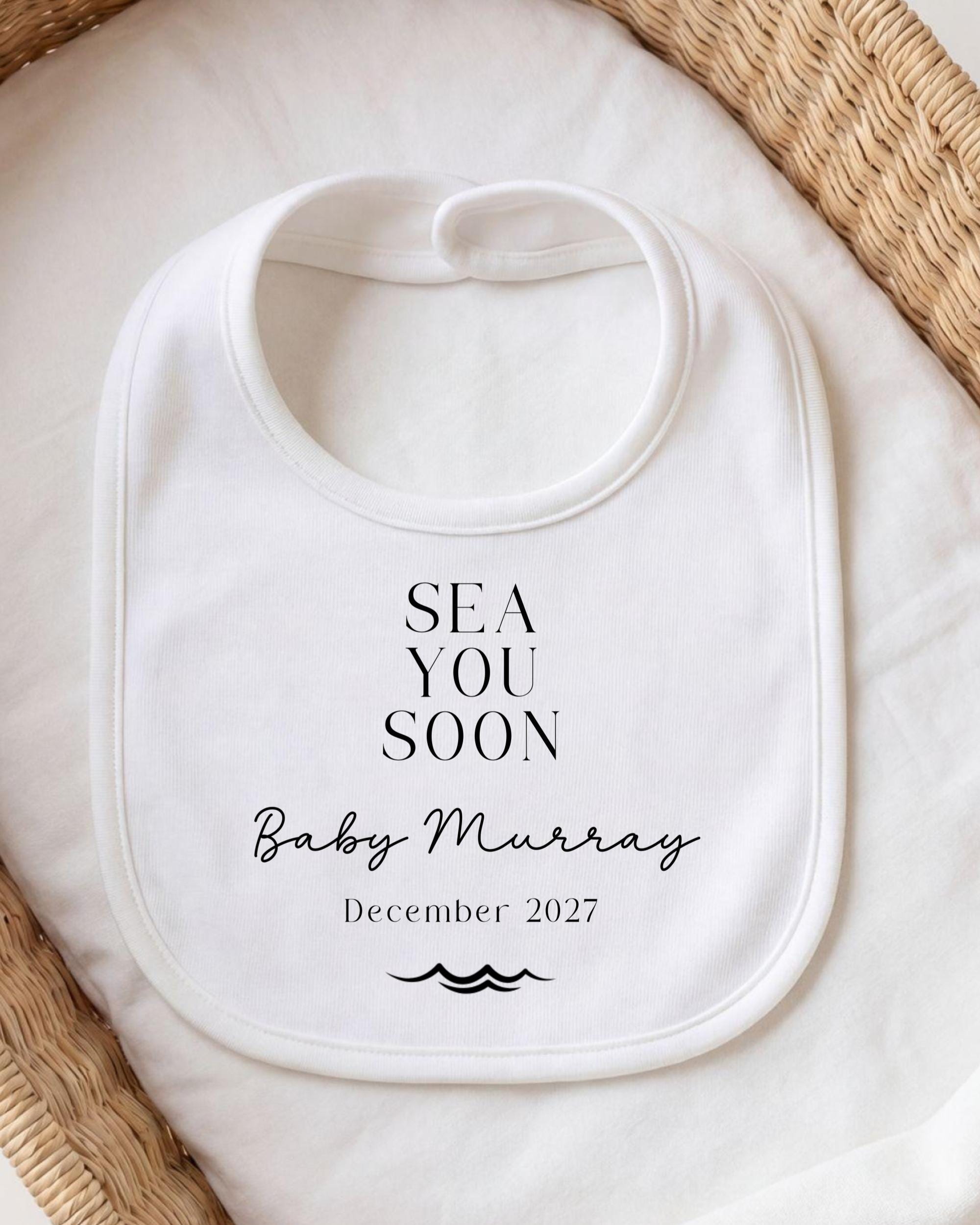 A white bib with the text "SEA YOU SOON Baby Murray December 2027" printed on it, placed on a white cloth.
