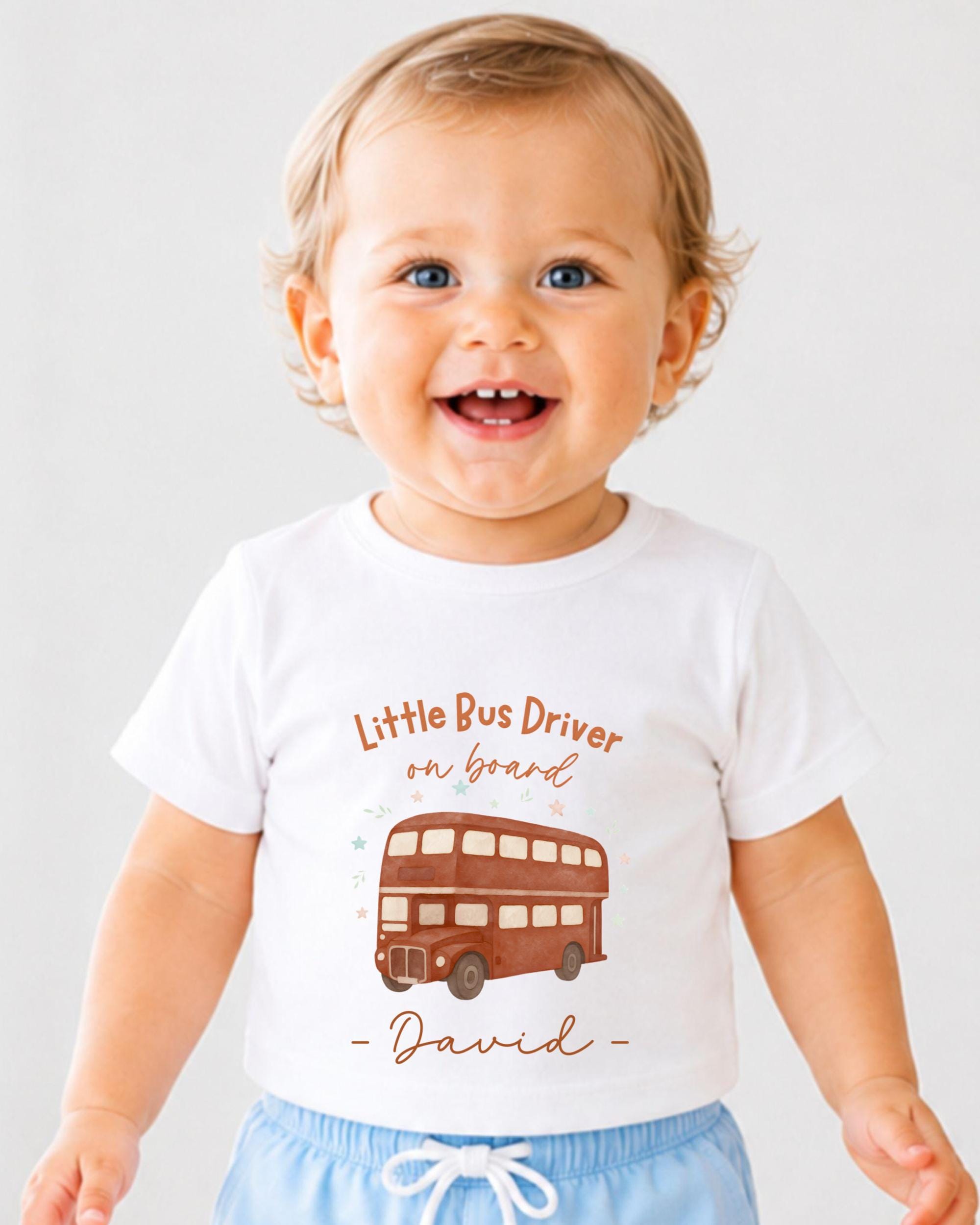 A smiling baby boy wearing a white t-shirt with a double-decker bus graphic and the text "Little Bus Driver on board David" printed on it.