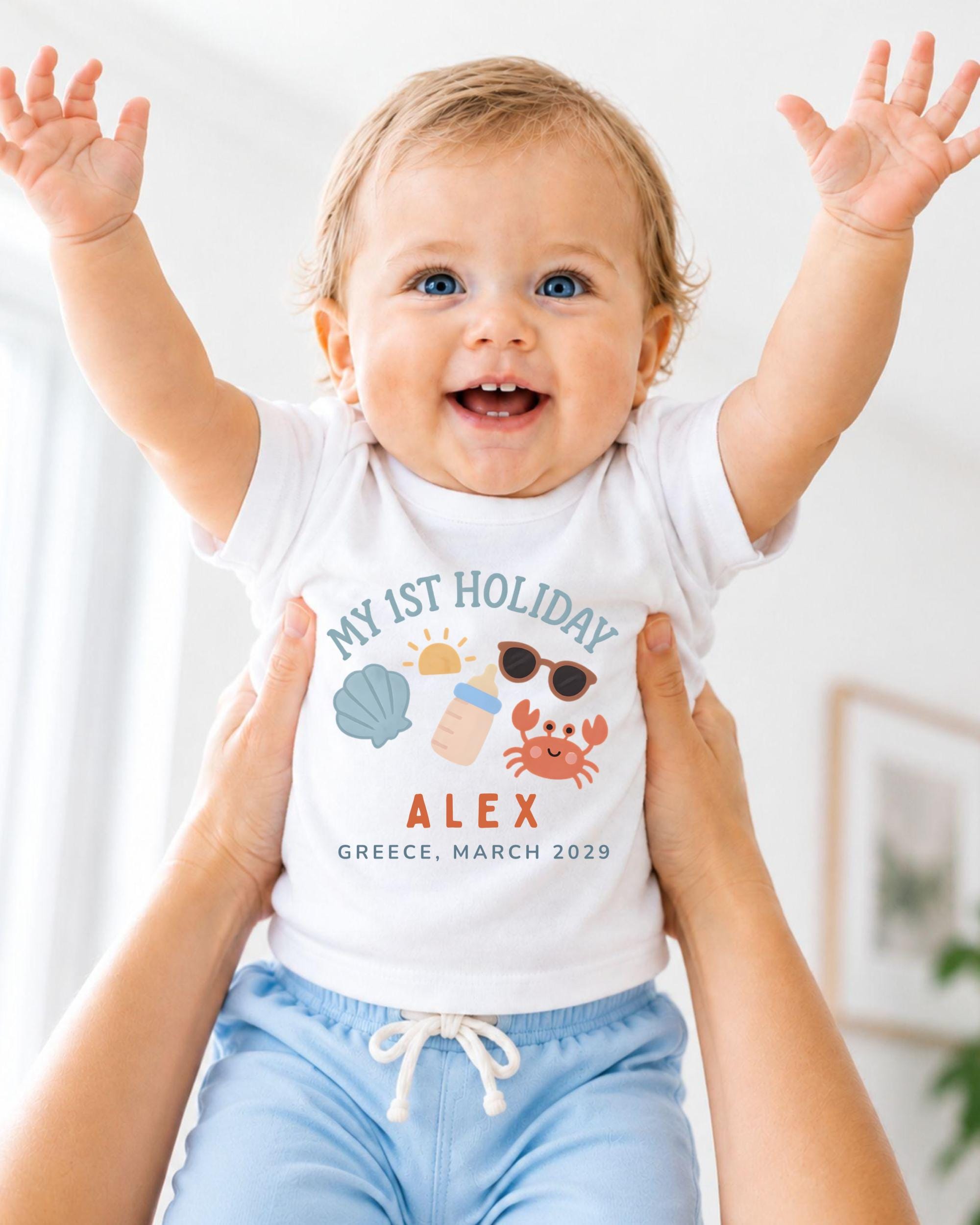A smiling baby wearing a white t-shirt with a personalized design and text, being held up by someone's arms.