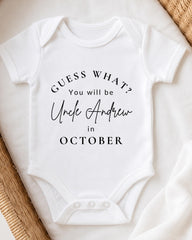 A white baby onesie with text printed on it, placed on a bed with a woven blanket.