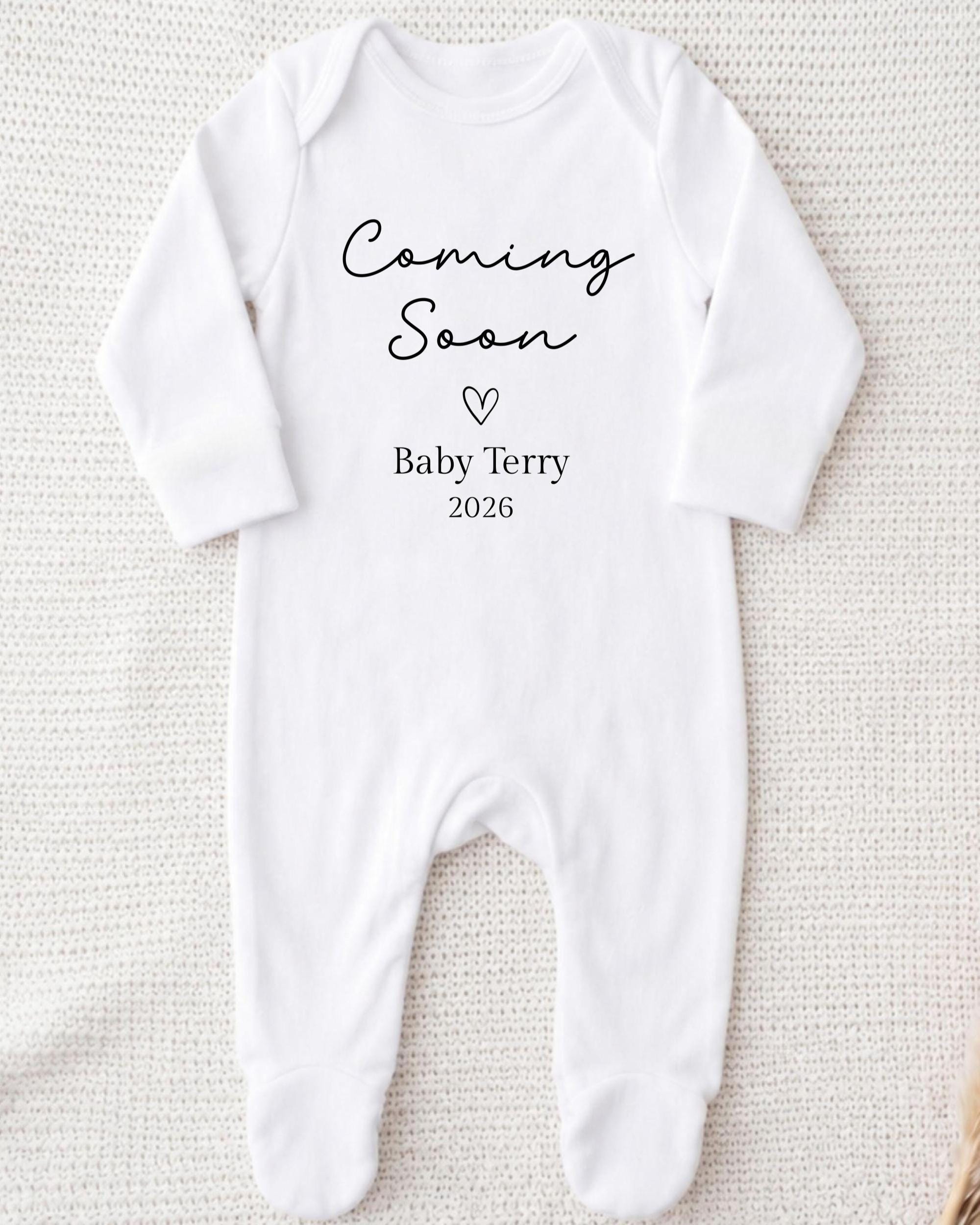 A white onesie with the text "Coming Soon Baby Terry 2026" printed on it.