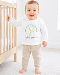 A smiling baby boy wearing a white sweatshirt with a tennis racket and the text "Born to play tennis with Daddy" is standing next to a wooden crib.
