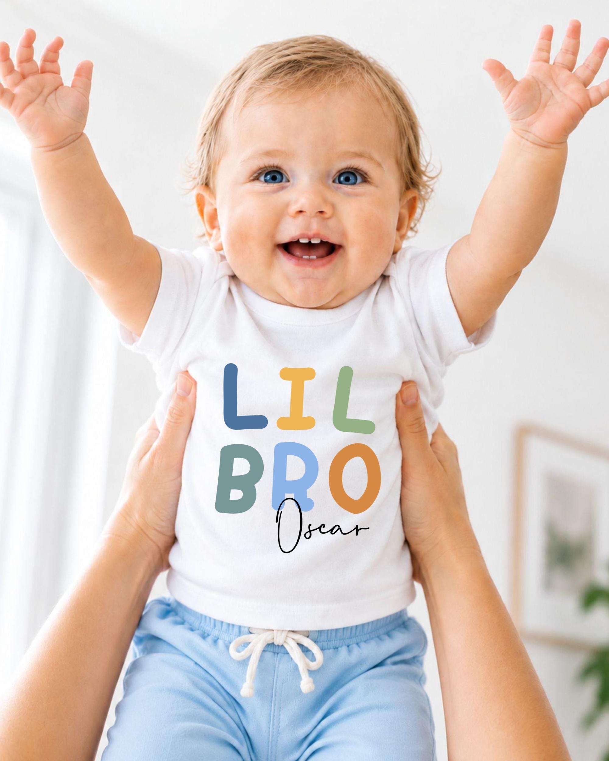 A smiling baby wearing a white t-shirt with the text "LIL BRO Oscar" is being held up by someone's arms.