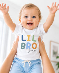 A smiling baby wearing a white t-shirt with the text "LIL BRO Oscar" is being held up by someone's arms.