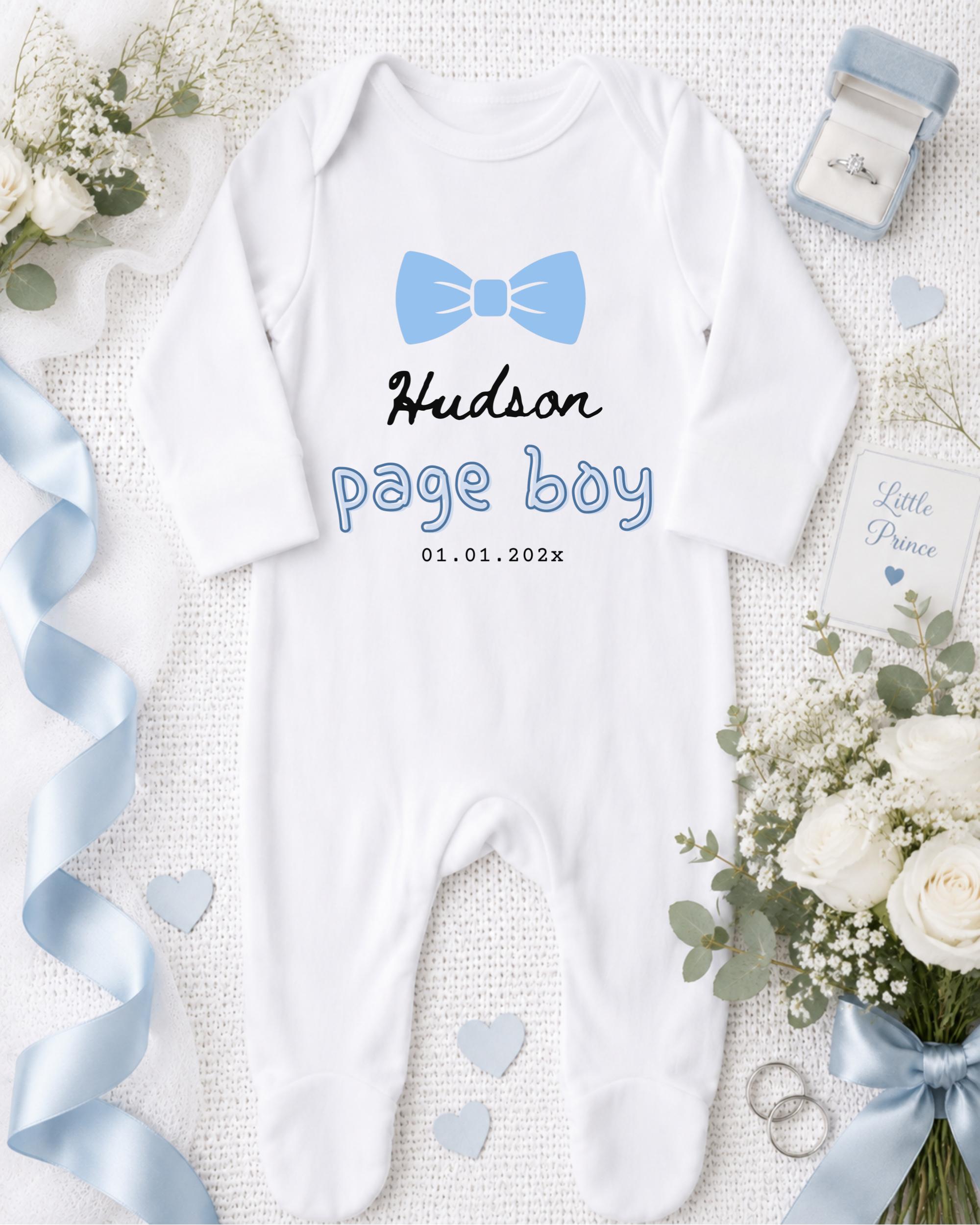 A white onesie with a blue bow tie, a ring, and a bouquet of white flowers are arranged on a white background.