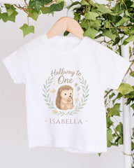 A white t-shirt with a hedgehog graphic and the text "Halfway to One - Izzy" printed on it, hanging on a wooden hanger.