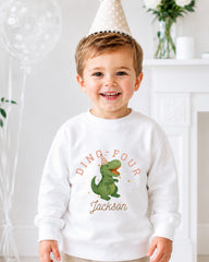 A young boy wearing a white sweatshirt with a dinosaur graphic and the text "DINO-FOUR JACKSON" is smiling at the camera.