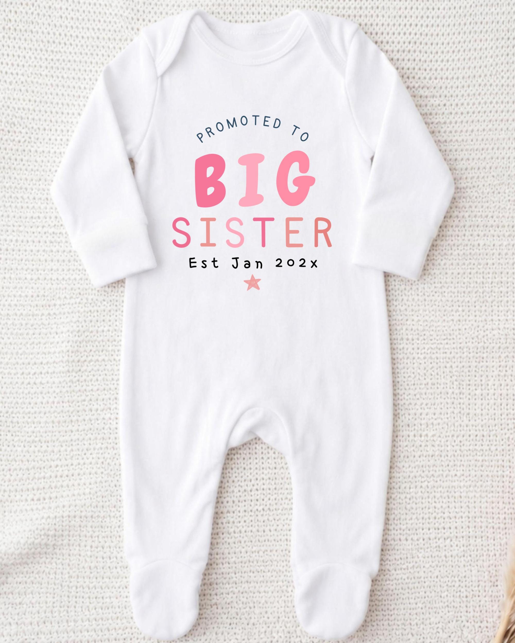 A white onesie with text that reads "Promoted to BIG SISTER Est Jan 2023*" printed on it.