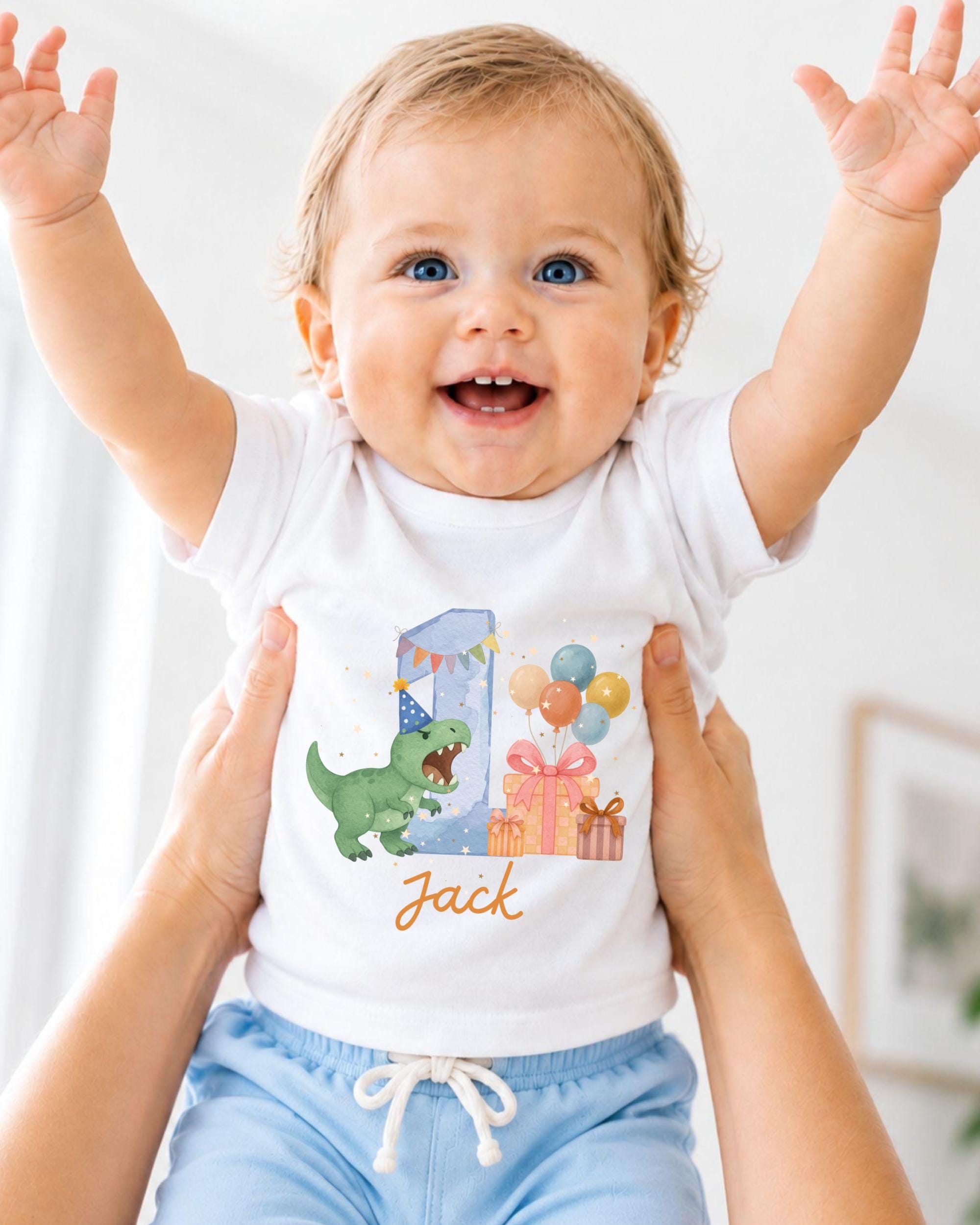A smiling baby wearing a white t-shirt with a dinosaur and birthday decorations, being held up by someone's arms.
