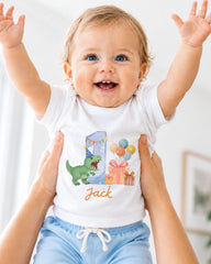A smiling baby wearing a white t-shirt with a dinosaur and birthday decorations, being held up by someone's arms.