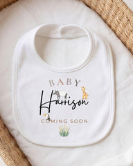 A white baby bib with the text "BABY Harrison COMING SOON" printed on it, along with illustrations of animals.
