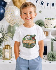 A young boy wearing a white t-shirt with a dinosaur graphic and the number 6, standing in front of a birthday party setup with balloons, a cake, and decorations.