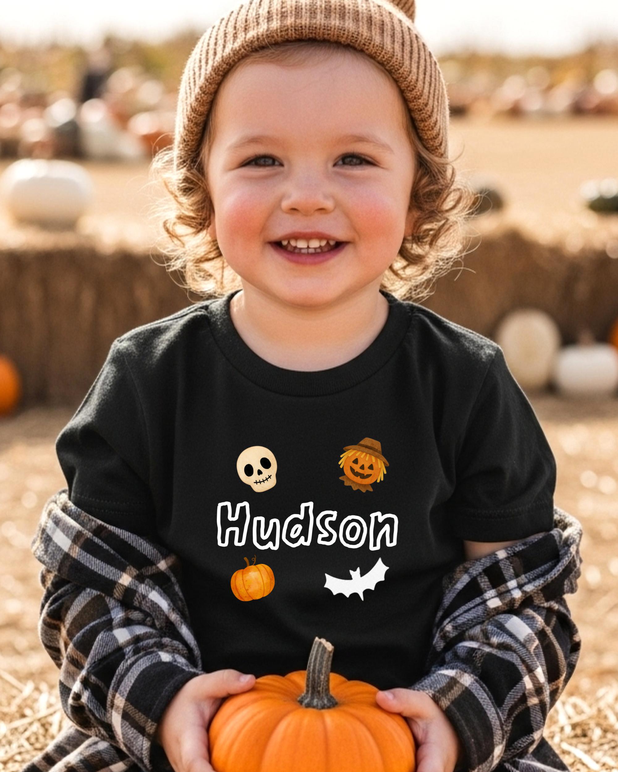 A young child wearing a black t-shirt with a pumpkin and bat design, smiling and holding a small orange pumpkin.