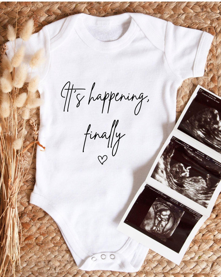 White Baby Vest with black script 'It's happening, finally' and an outline heart for a IVF pregnancy announcement.