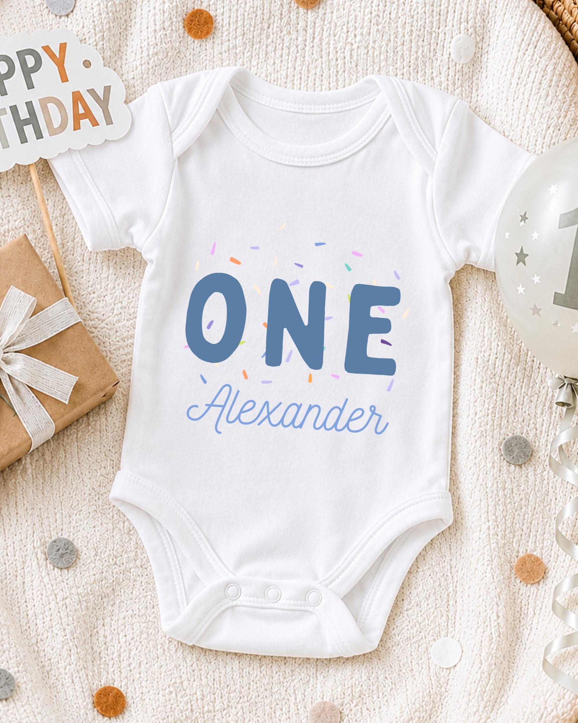 A white onesie with the text "ONE Alexander" printed on it, surrounded by confetti and a birthday sign.