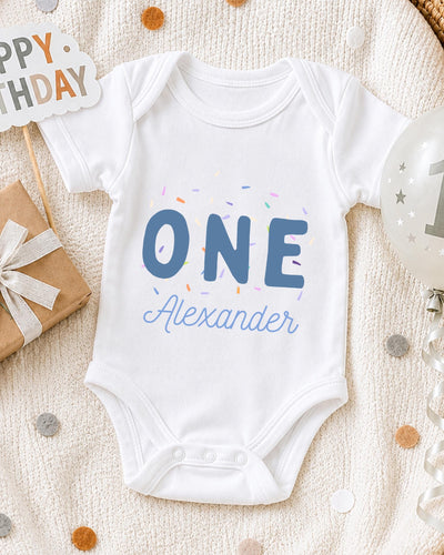 A white bodysuit with the text "ONE Alexander" printed on it, surrounded by confetti and a birthday sign.