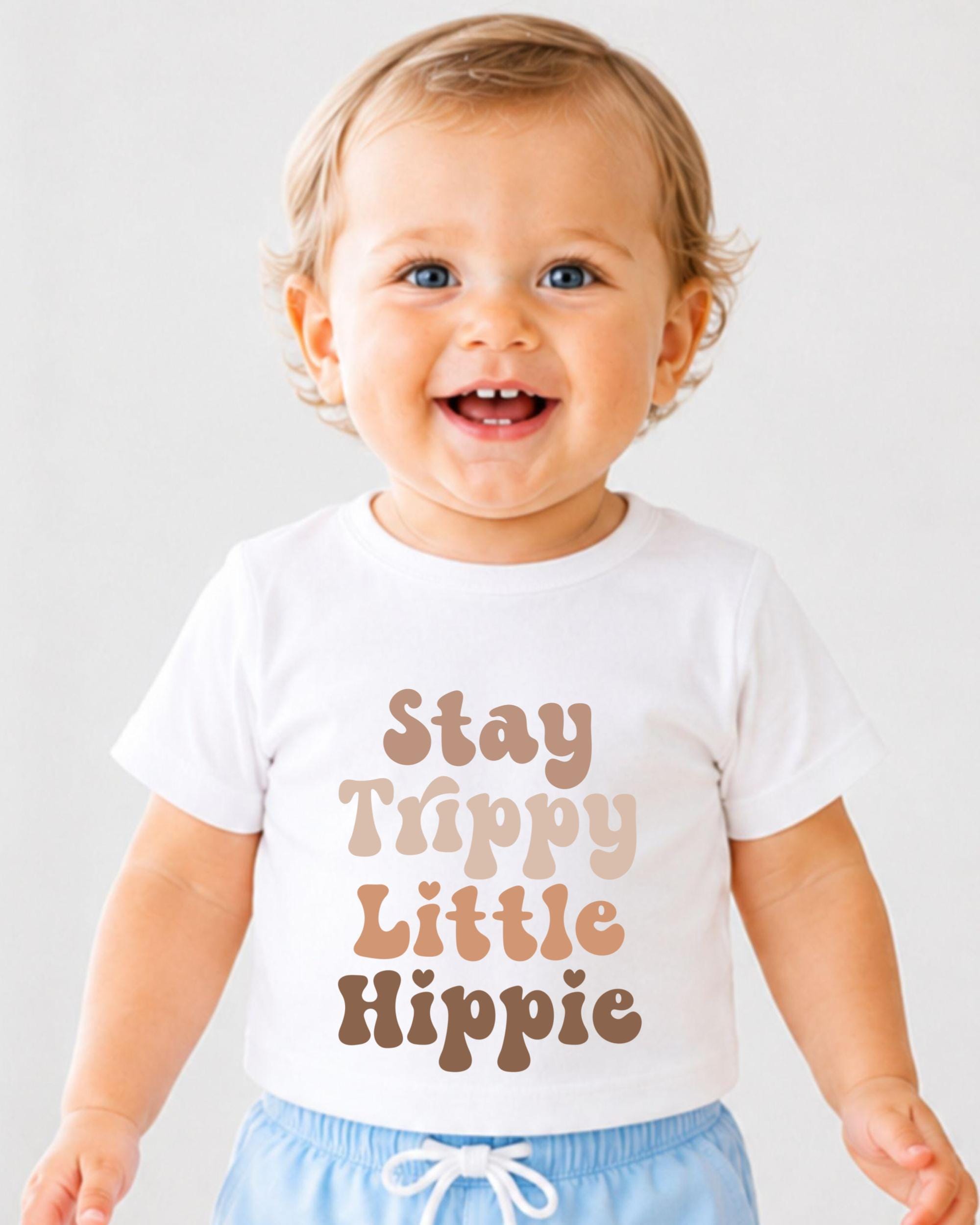 A smiling baby wearing a white t-shirt with the text "Stay Trippy Little Hippie" printed on it.