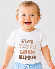 A smiling baby wearing a white t-shirt with the text "Stay Trippy Little Hippie" printed on it.