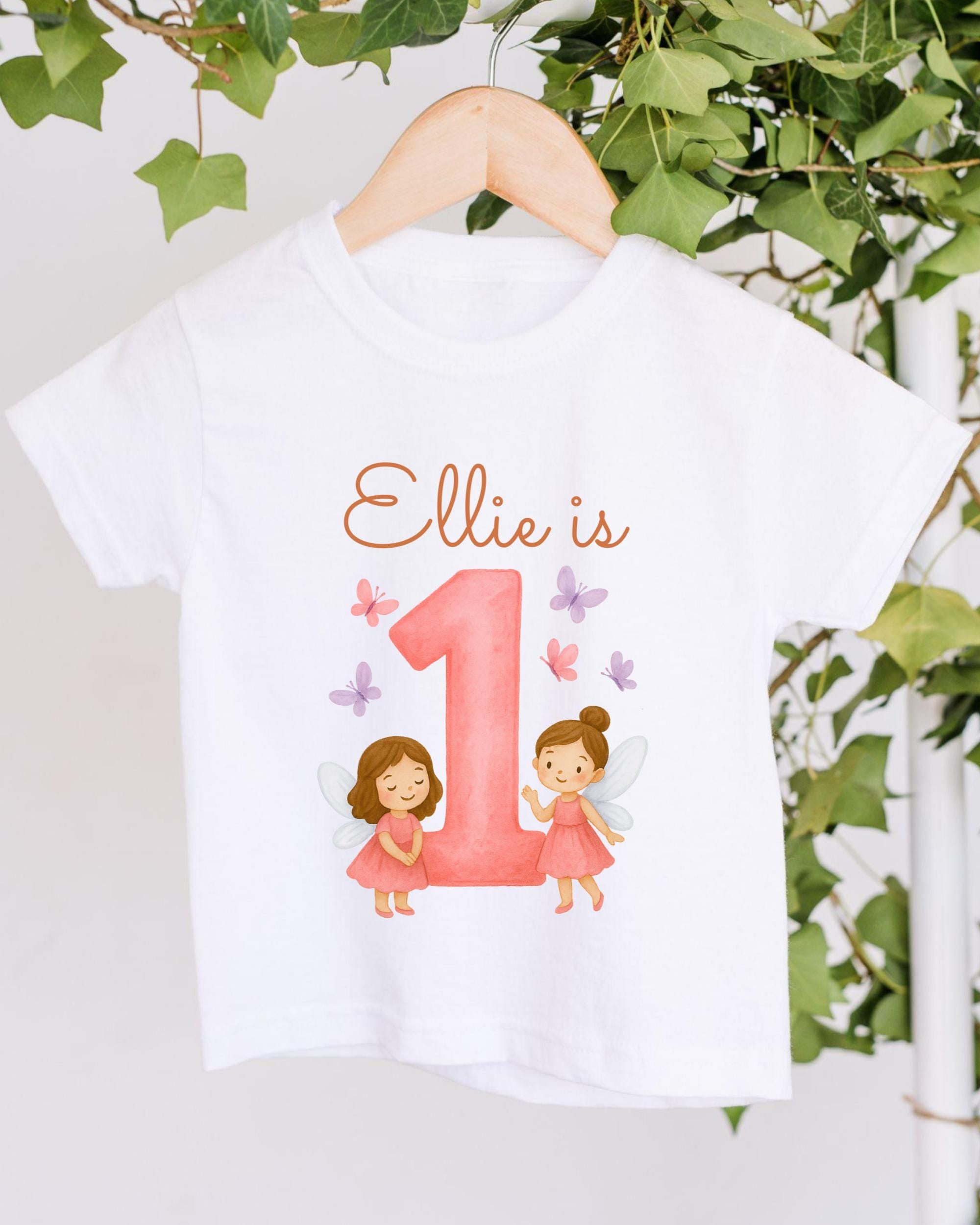 A white t-shirt with a pink number one and two-legged fairy characters printed on it, along with the text "Ellie is 1".