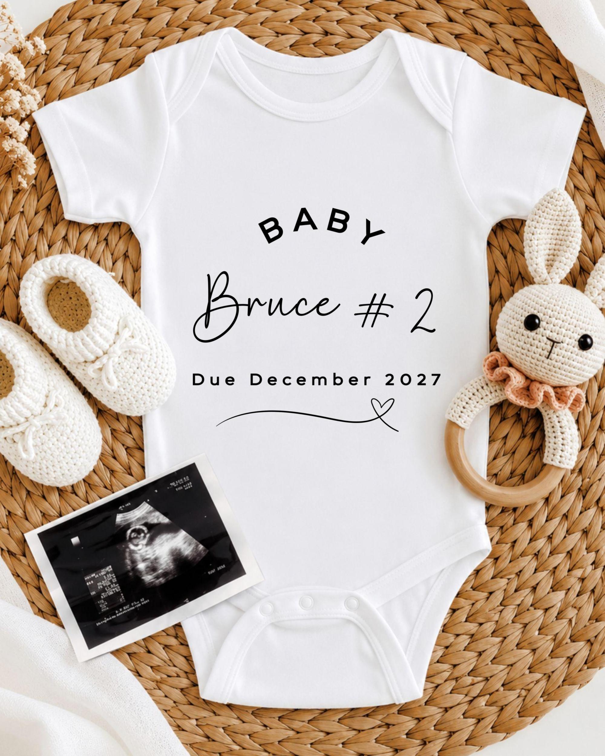 A white baby onesie with the text "BABY Bruce #2 Due December 2027" printed on it, along with a crocheted rabbit toy and a pair of white booties. There is also an Ultrasound image of a baby's belly.