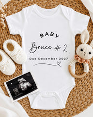 A white baby onesie with the text "BABY Bruce #2 Due December 2027" printed on it, along with a crocheted rabbit toy and a pair of white booties. There is also an Ultrasound image of a baby's belly.