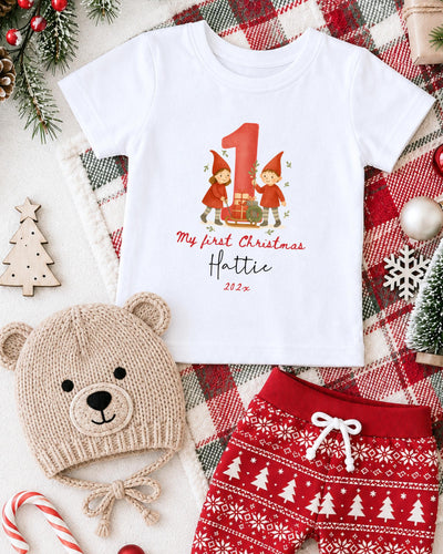A white t-shirt with a graphic of two gnomes and the text "My first Christmas" printed on it, along with a teddy bear hat and red shorts, all set against a Christmas-themed background.