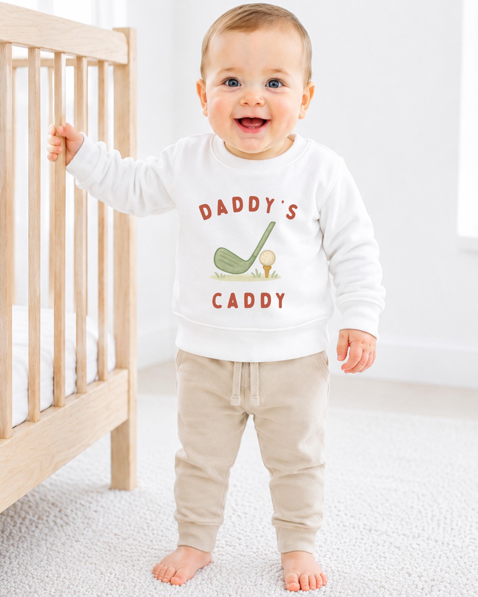 A smiling baby boy wearing a white sweatshirt with the text "DADDY'S CADDY" printed on it, standing next to a wooden crib.