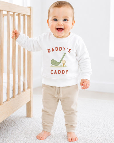 A smiling baby boy wearing a white sweatshirt with the text "DADDY'S CADDY" printed on it, standing next to a wooden crib.