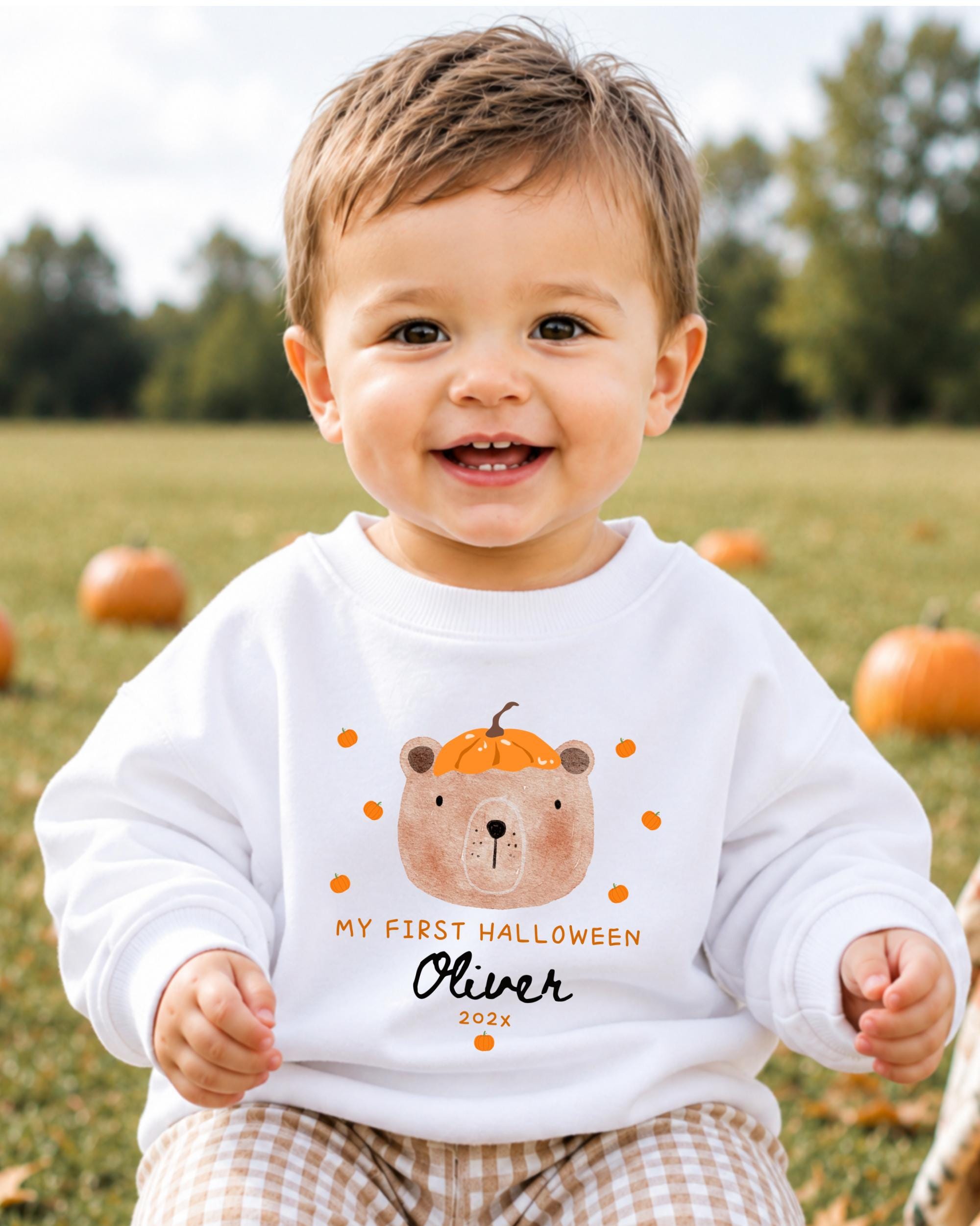 A smiling baby boy wearing a white sweatshirt with a pumpkin and bear design, sitting in a field with pumpkins in the background.