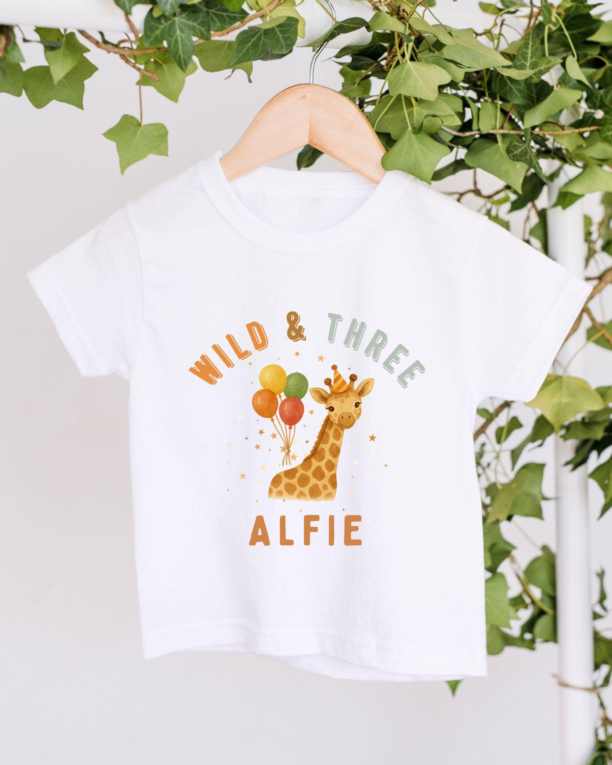 A white t-shirt with a giraffe and balloons printed on it, hanging on a wooden hanger.