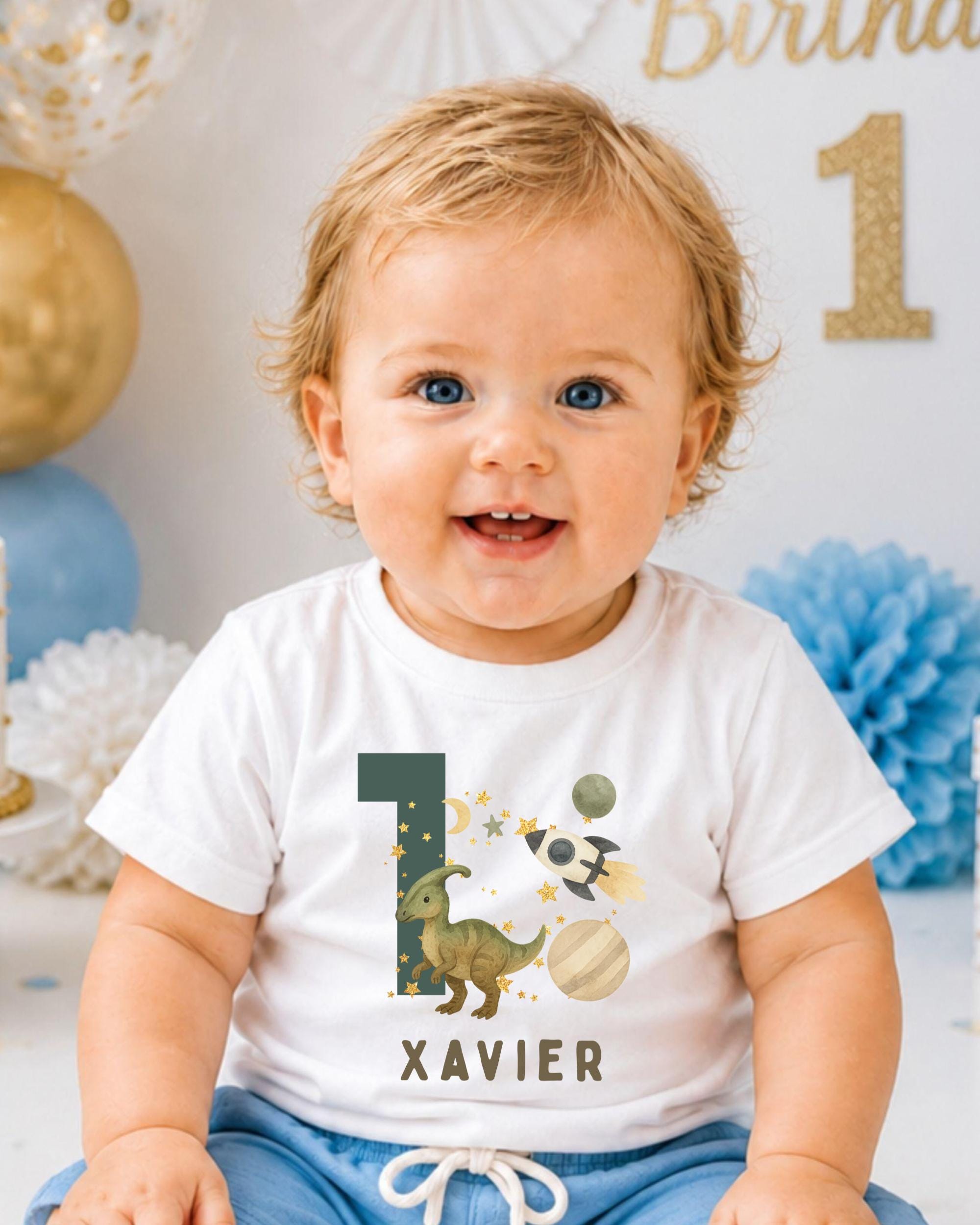 A smiling baby boy wearing a white t-shirt with a dinosaur graphic and the number 1, sitting in front of a birthday party backdrop.