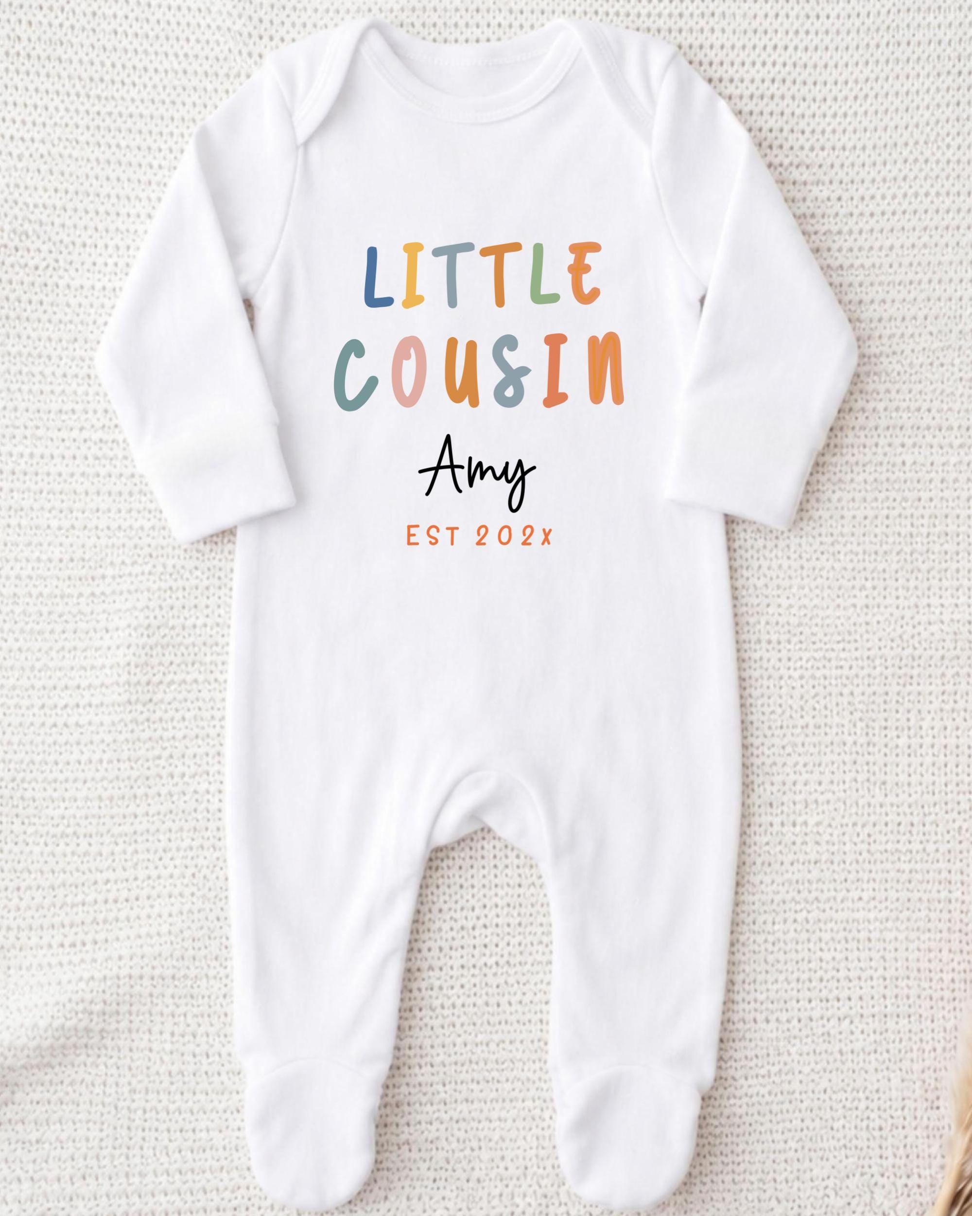A white onesie with the text "LITTLE COUSIN Amy EST 202x" printed on it.