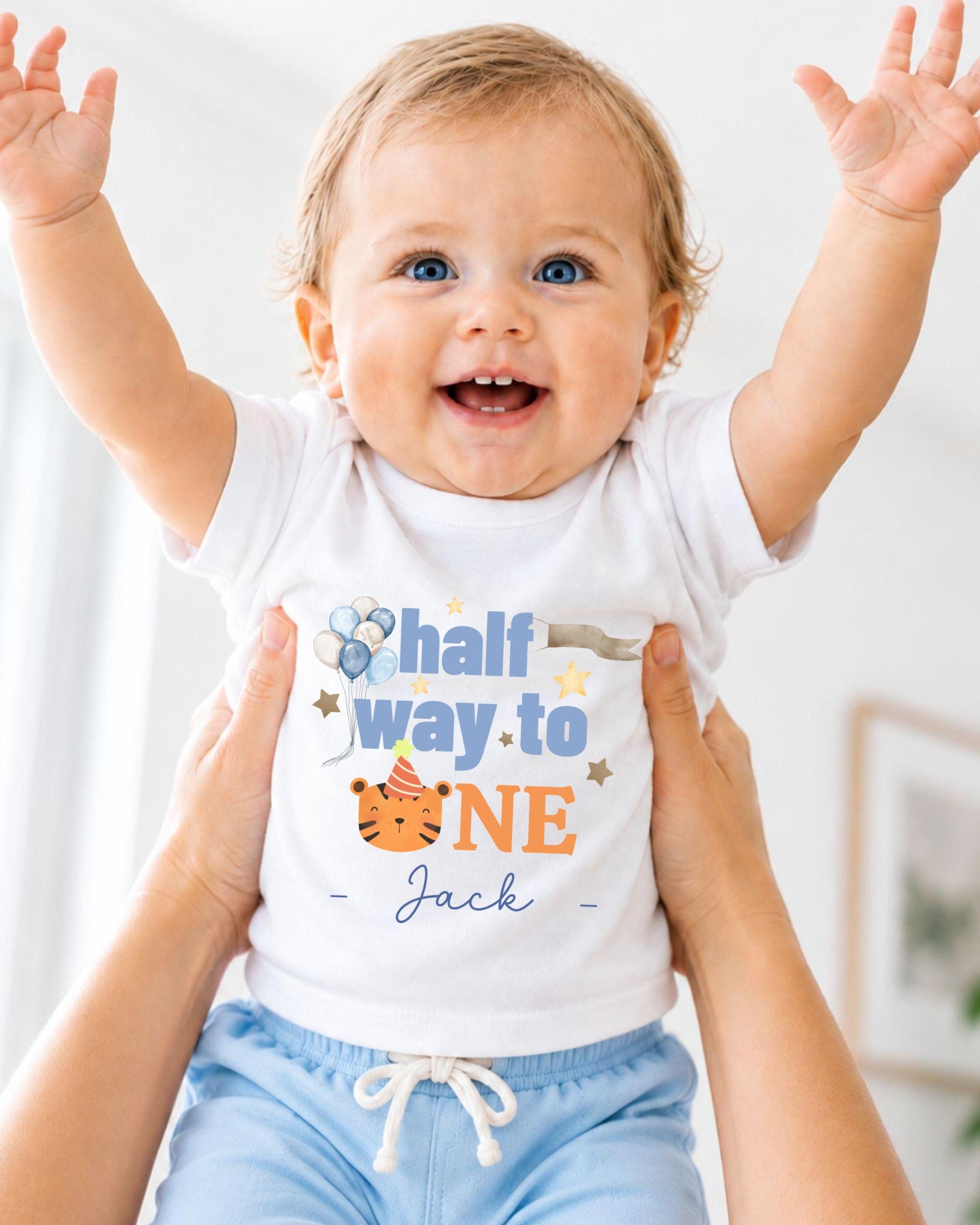 A smiling baby wearing a white t-shirt with a birthday message and a tiger graphic, being held up by someone's arms.