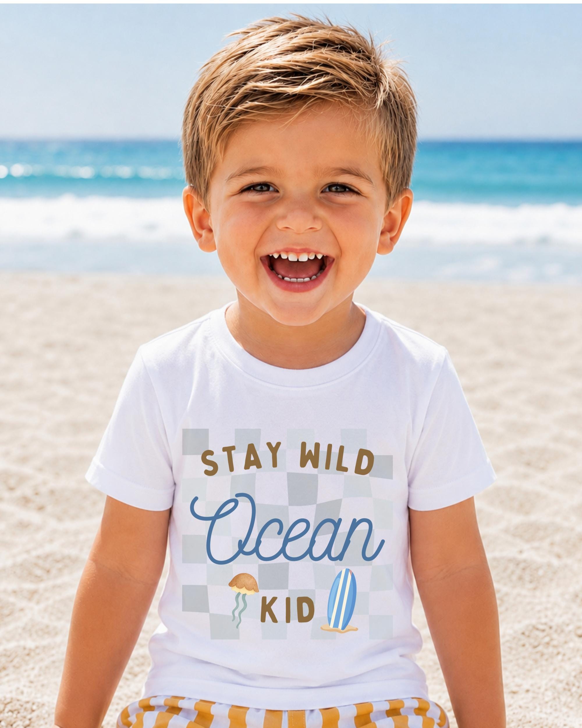 A young boy with a big smile on his face, wearing a white t-shirt with the text "STAY WILD OCEAN KID" printed on it, is standing on a sandy beach with the ocean in the background.