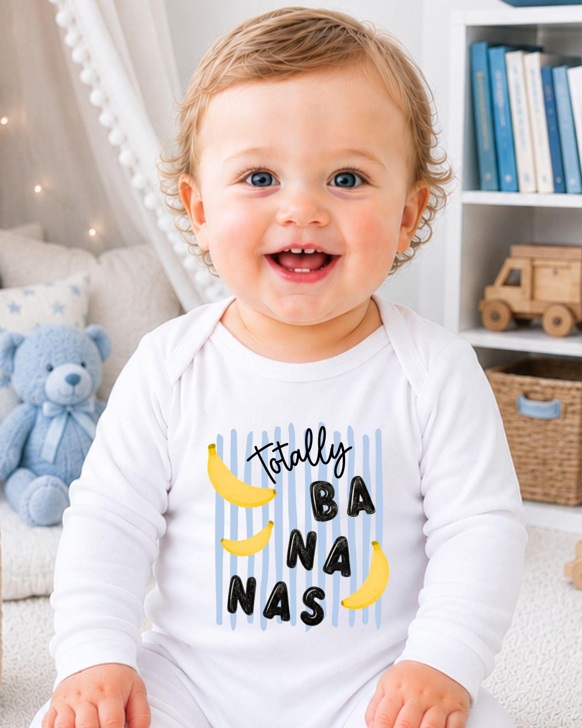 A smiling baby wearing a white onesie with a banana and text printed on it, sitting in a room with a teddy bear and bookshelf in the background.