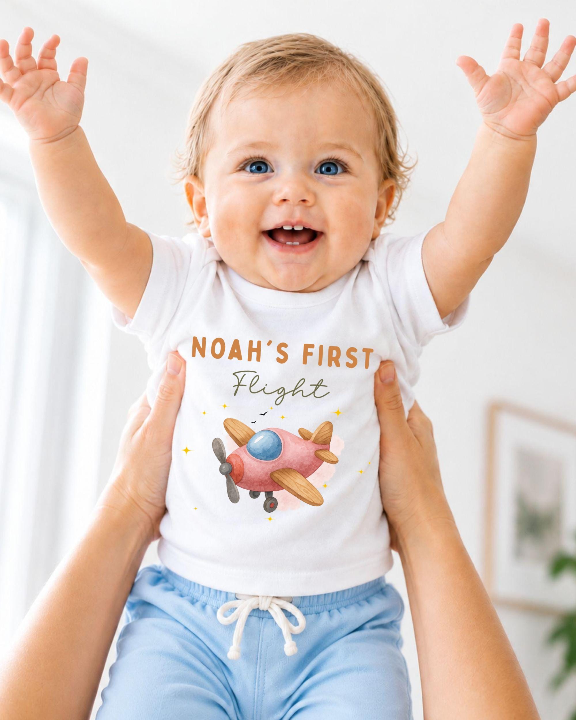 A smiling baby wearing a white t-shirt with a pink airplane graphic and the text "Noah's first flight" is being held up by two people.