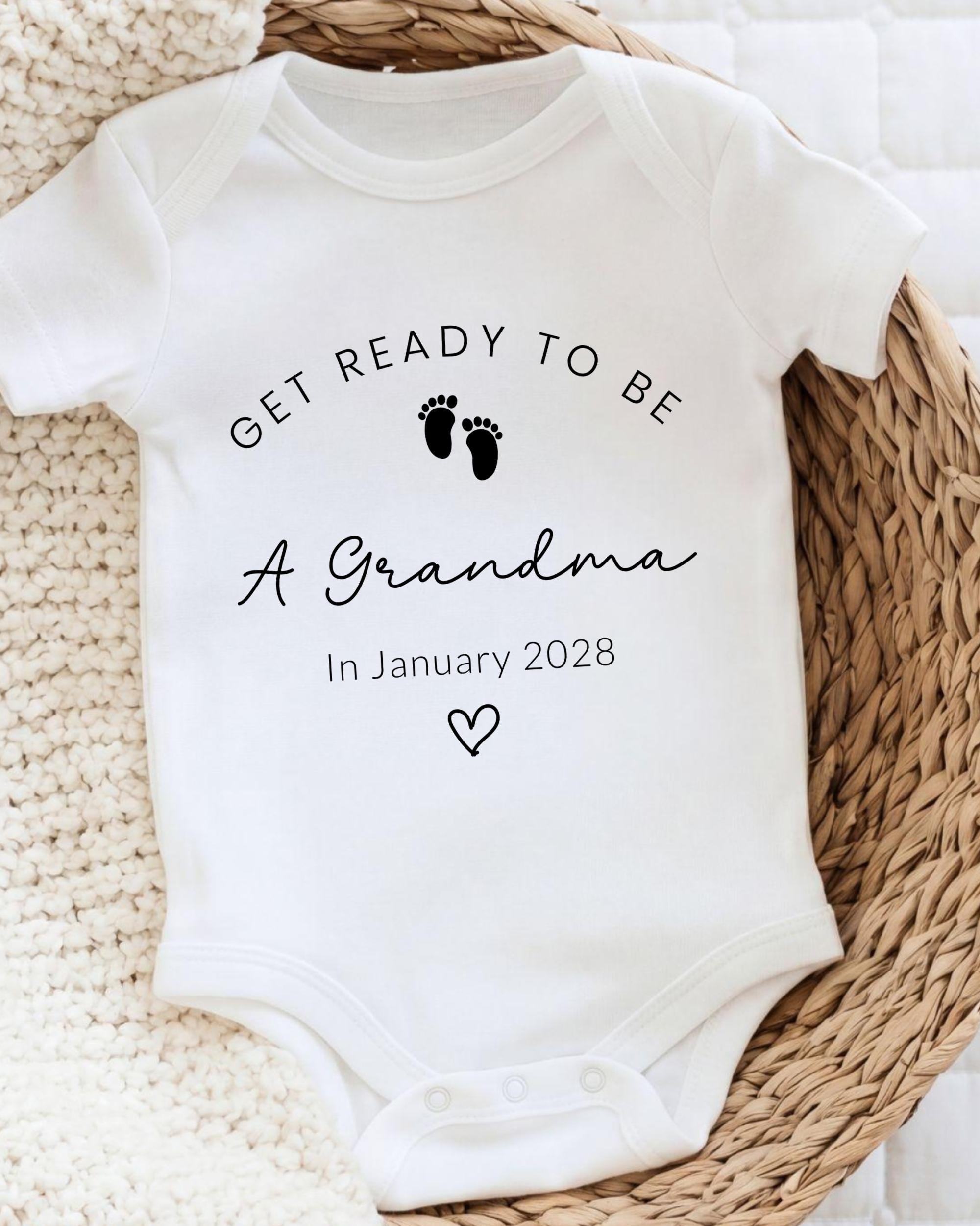 A white baby onesie with the text "GET READY TO BE A GRANDMA In January 2028" printed on it, along with a small heart symbol.