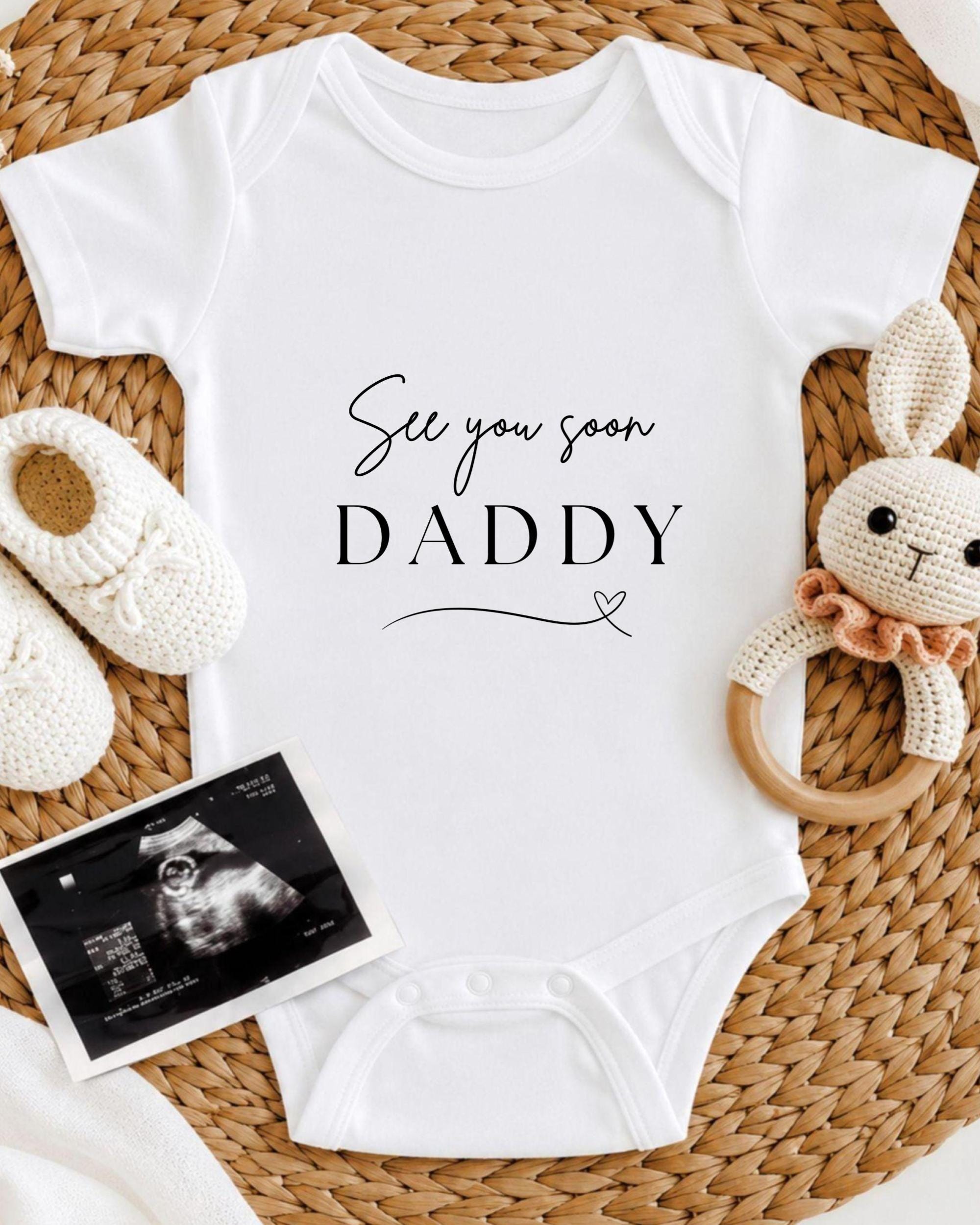 A white baby onesie with the text "See you soon Daddy" printed on it, along with a crocheted bunny toy and a black and white photograph of an infant's head.