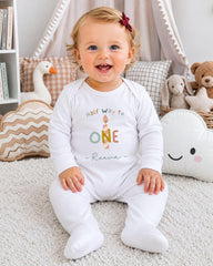 A smiling baby girl wearing a white onesie with the text "Half Way to One" sits on a white carpet surrounded by stuffed animals and pillows.