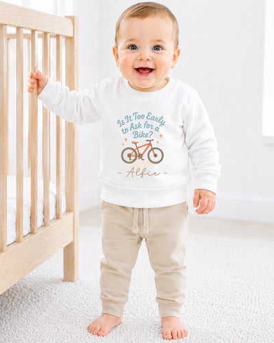 A smiling baby boy wearing a white sweatshirt with a bicycle graphic and the text "Is it too early to ask for a bike?"